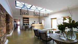 A spacious room with a high ceiling, a stone wall, a wooden reception desk, a dining area with chairs and a table, and two large hanging light fixtures. - The Old Rectory (Plettenberg Bay)
