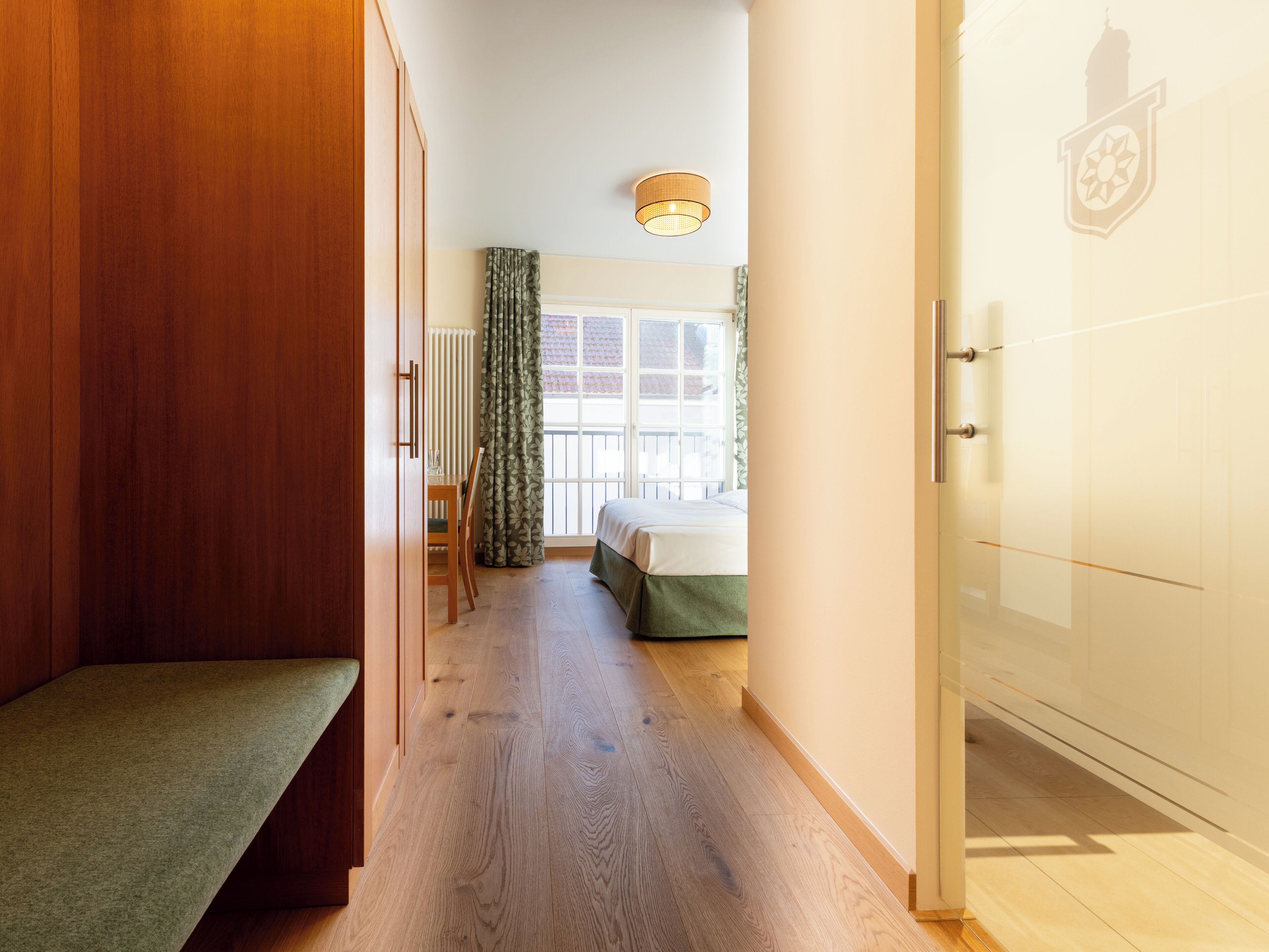 A hallway with a wooden bench, a bed with a green bedspread, a window with curtains, and a radiator.