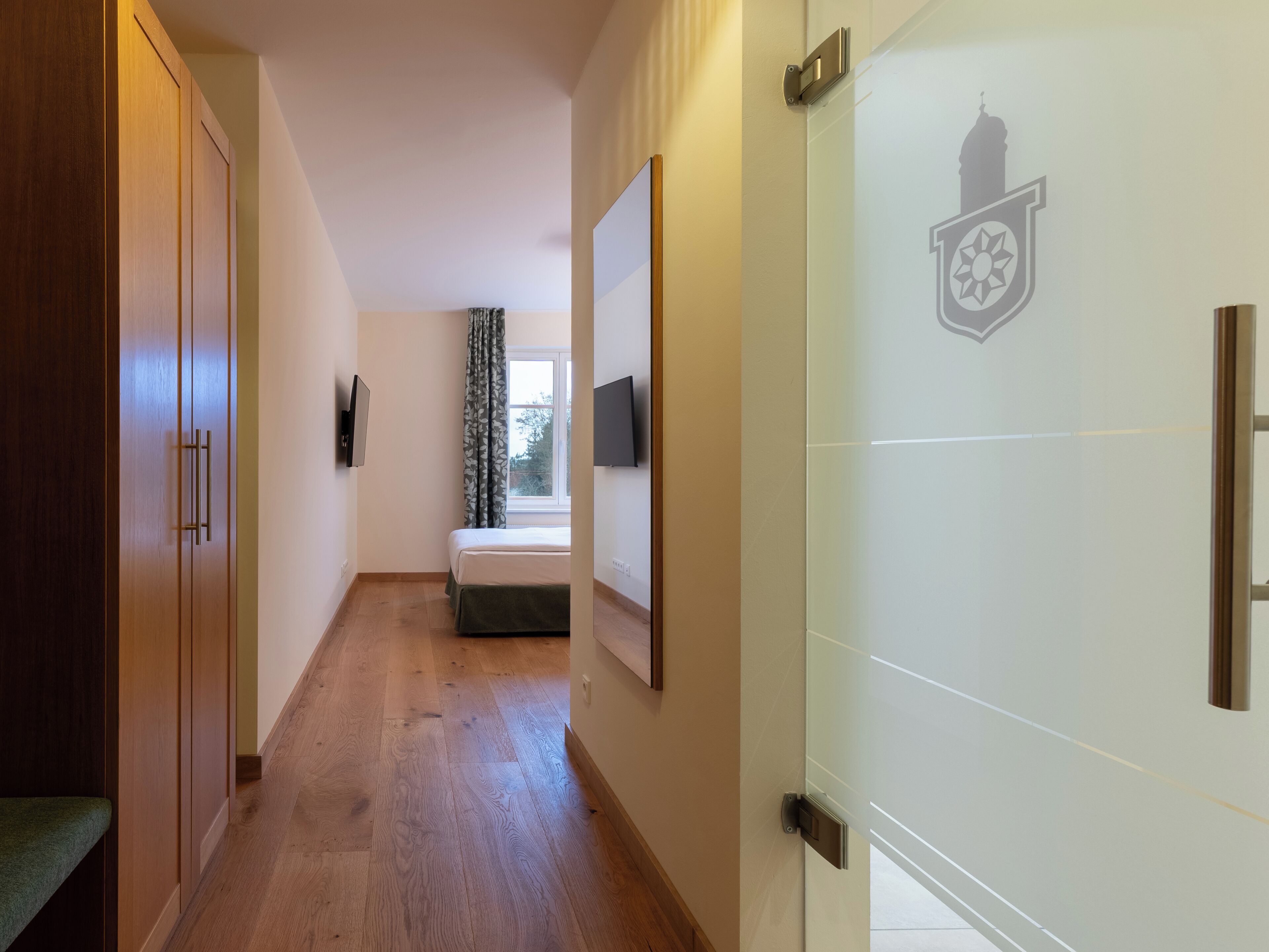 Un pasillo con piso de madera, una cama con colcha verde, una ventana con vista a árboles y una puerta con un escudo de armas.