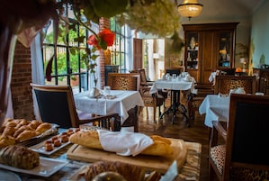 Restaurante com várias mesas, cadeiras e uma variedade de doces e pães expostos num balcão.