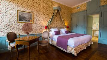 A bedroom with a large bed, a wooden desk, two chairs, a chandelier, and floral wallpaper.