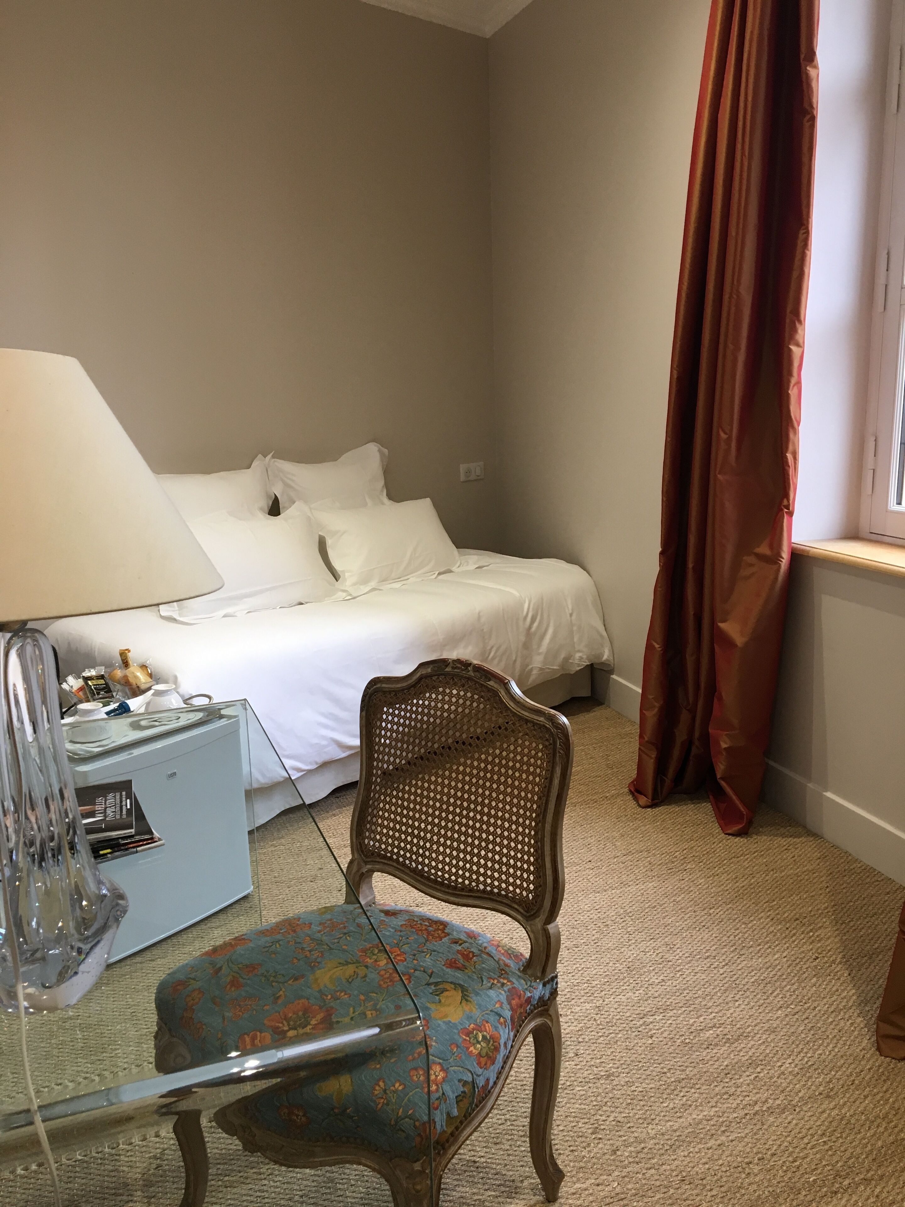 A hotel room with a bed, a desk with a lamp, a chair, and a window with curtains.