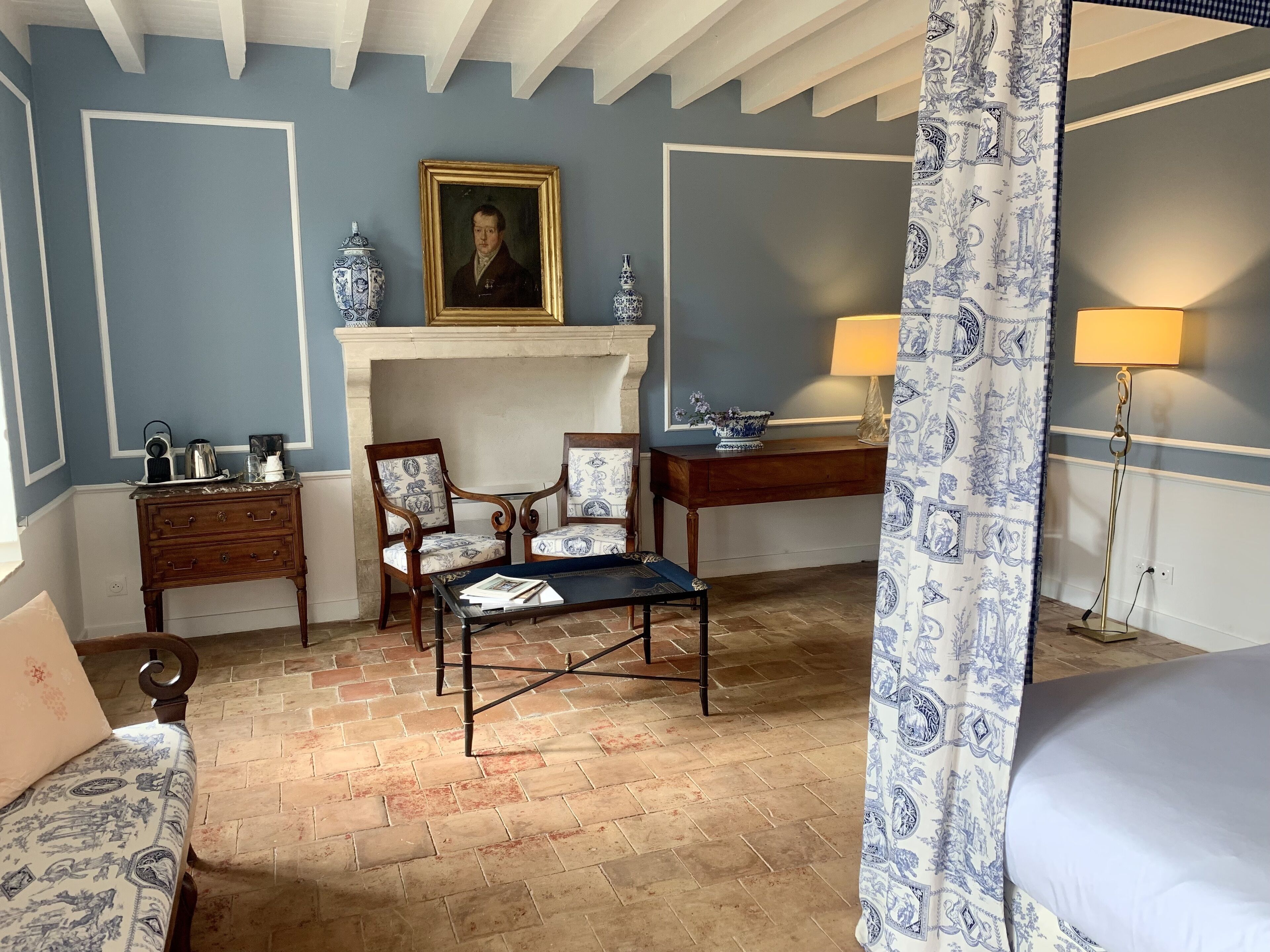 Una habitación con chimenea, dos sillones, una mesita, una cama y un escritorio con lámpara.