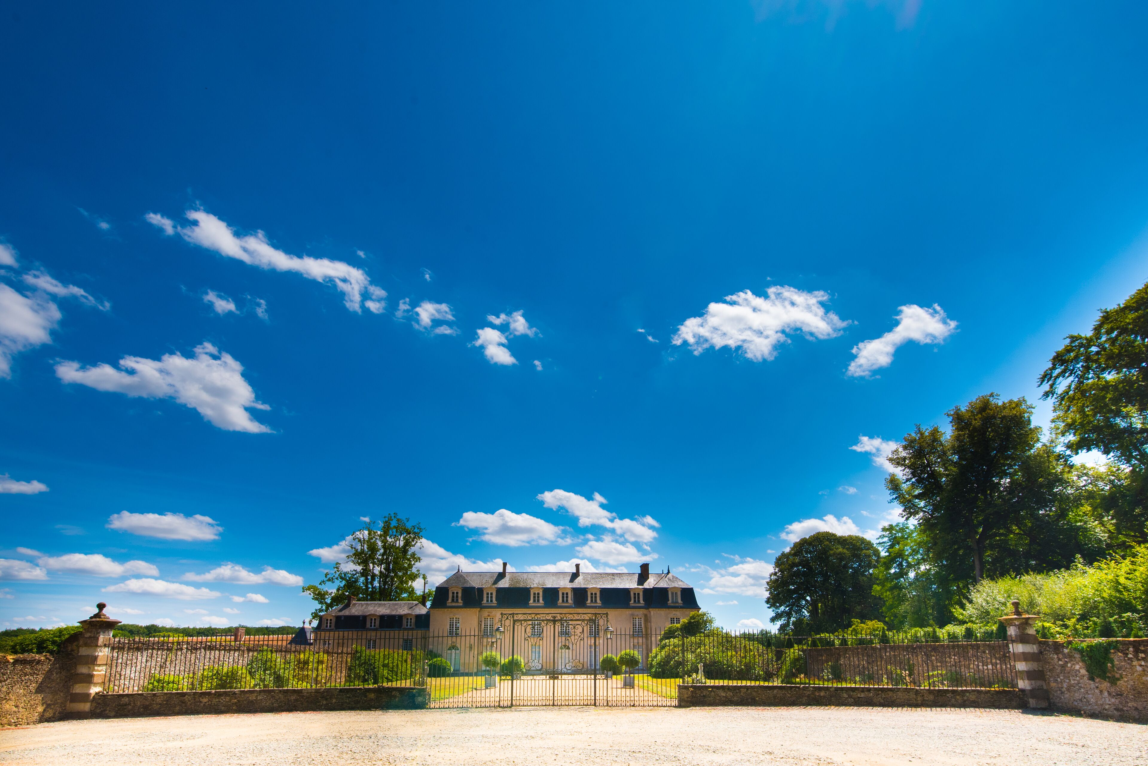 A large, historic building with a symmetrical facade, surrounded by a well-maintained garden and a gravel driveway.
