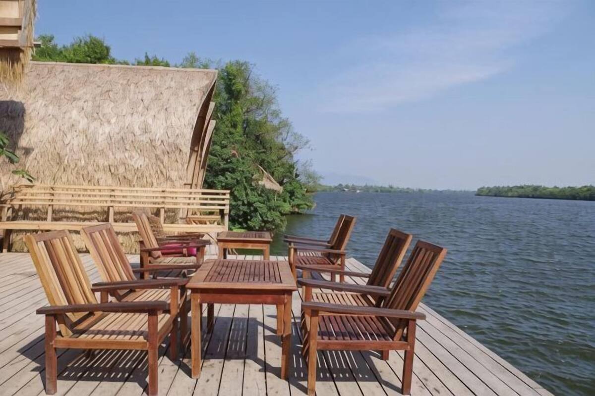 Une terrasse en bois, agrémentée de chaises et de tables, qui offre une vue sur une étendue d’eau, ainsi qu’une structure à toit de chaume et de la verdure.