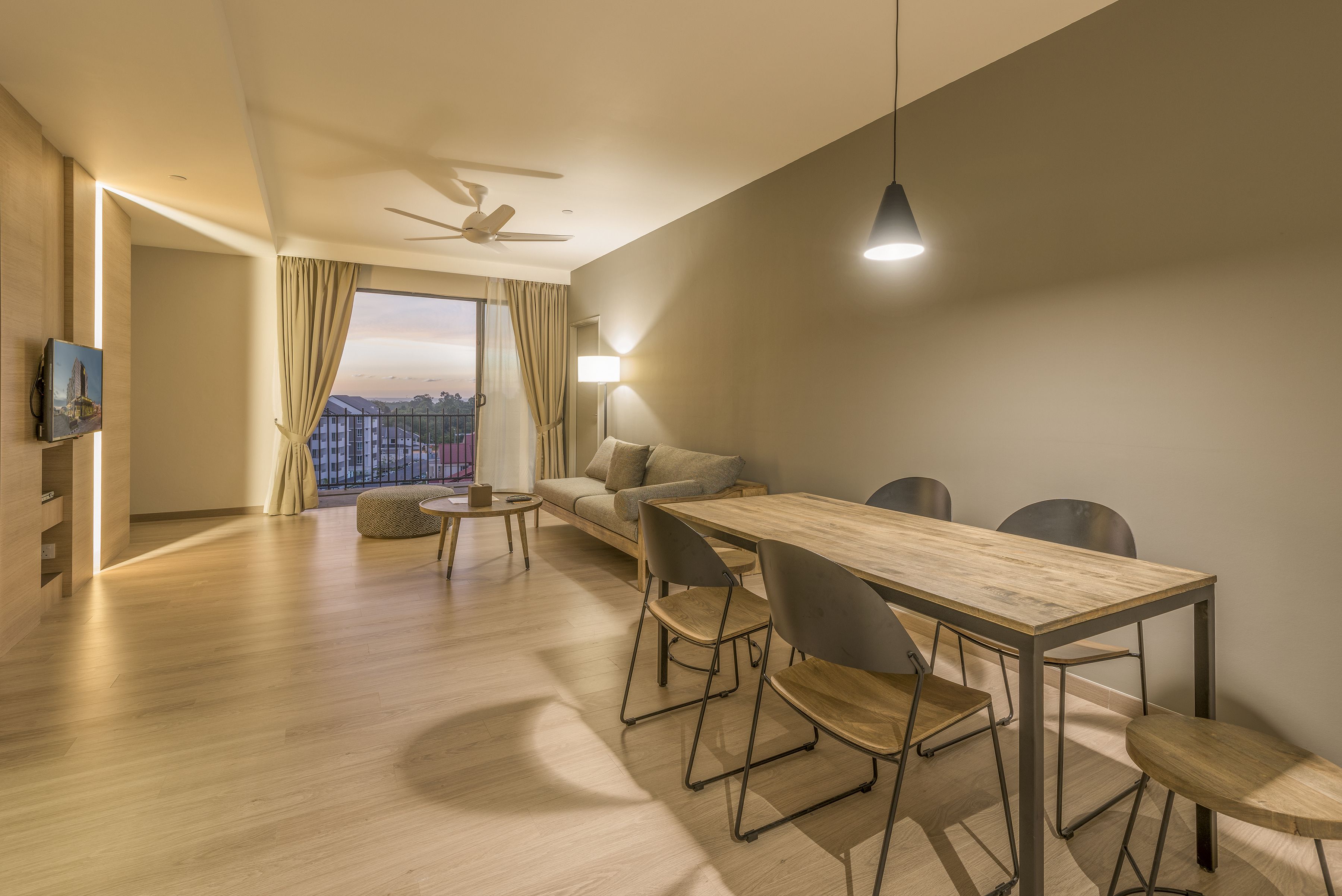 Una sala moderna con una mesa de comedor de madera, sillas negras, un sofá, un televisor montado en la pared y un amplio ventanal con cortinas.