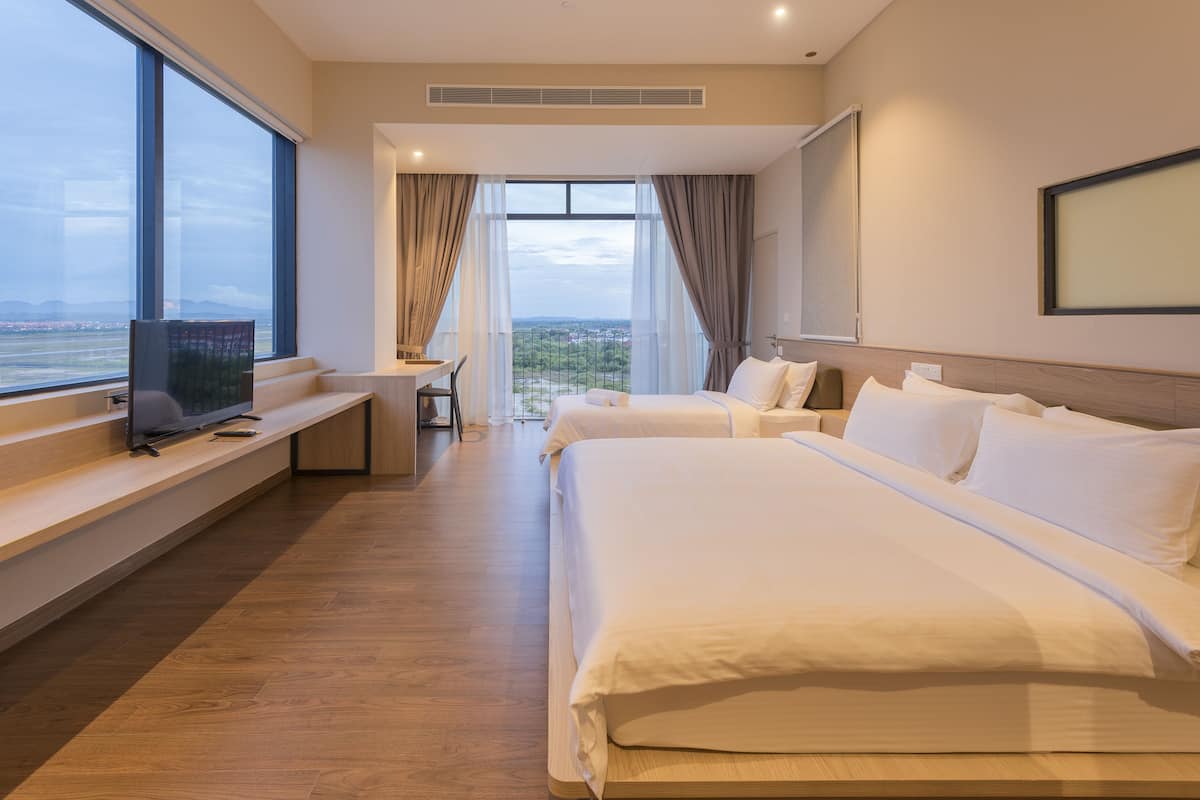 A hotel room with two beds, a TV on a bench, a desk, and large windows offering a view of the city.