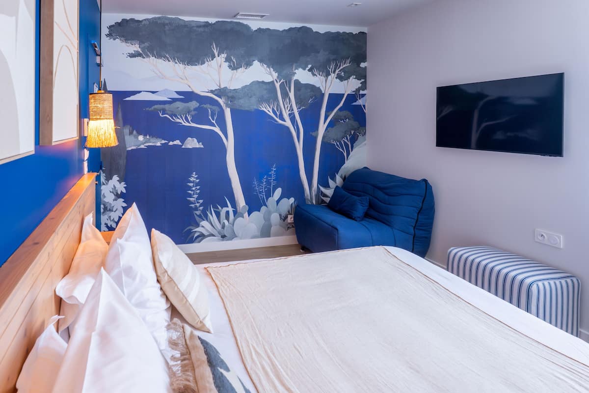 A bedroom with a bed, a blue chair, a striped ottoman, a wall-mounted TV, and a mural of trees.