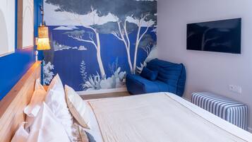 Une chambre à coucher avec un lit, une chaise bleue, un pouf à rayures, un téléviseur fixé au mur et une fresque représentant des arbres.