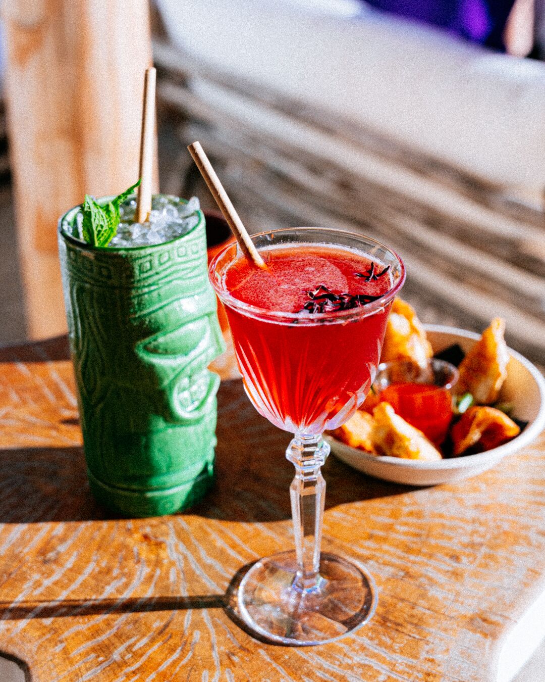 Um copo de cocktail com canudo e folha de hortelã, um copo verde com canudo, e uma tigela de comida sobre uma mesa de madeira.