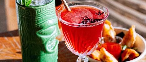 Um copo de cocktail com canudo e folha de hortelã, um copo verde com canudo, e uma tigela de comida sobre uma mesa de madeira.