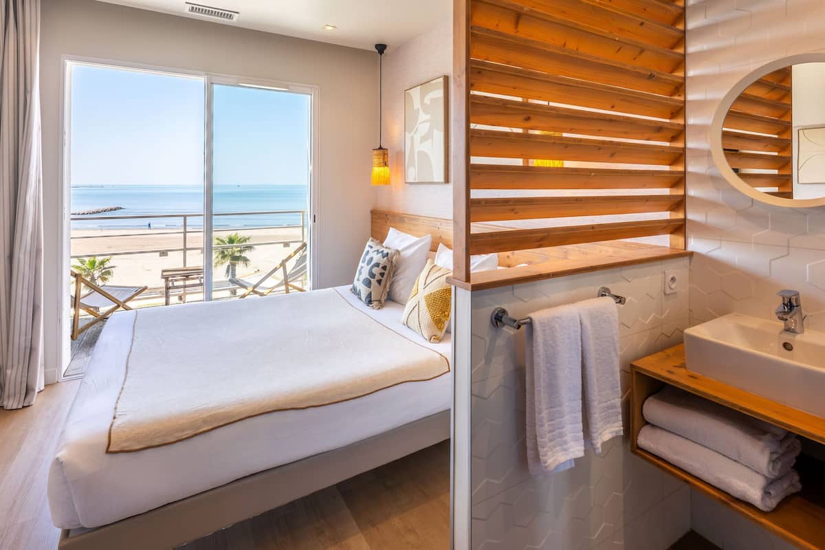 A bedroom with a large window, a wooden slatted wall, a bed, a bedside table, a sink, and a towel rack.