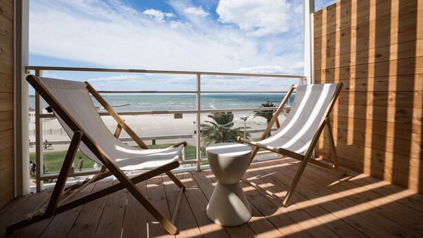 A balcony with two white cushioned chairs and a white table, overlooking a beach and ocean. - Hôtel Café Miramar (Le Grau-Du-Roi)