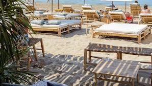 A beachside lounge area with wooden lounge chairs and umbrellas, a thatched roof, and a view of the sea. - Hôtel Café Miramar (Le Grau-Du-Roi)