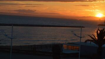 A sunset view from a balcony overlooking a calm sea, with a pier and a palm tree in the foreground.