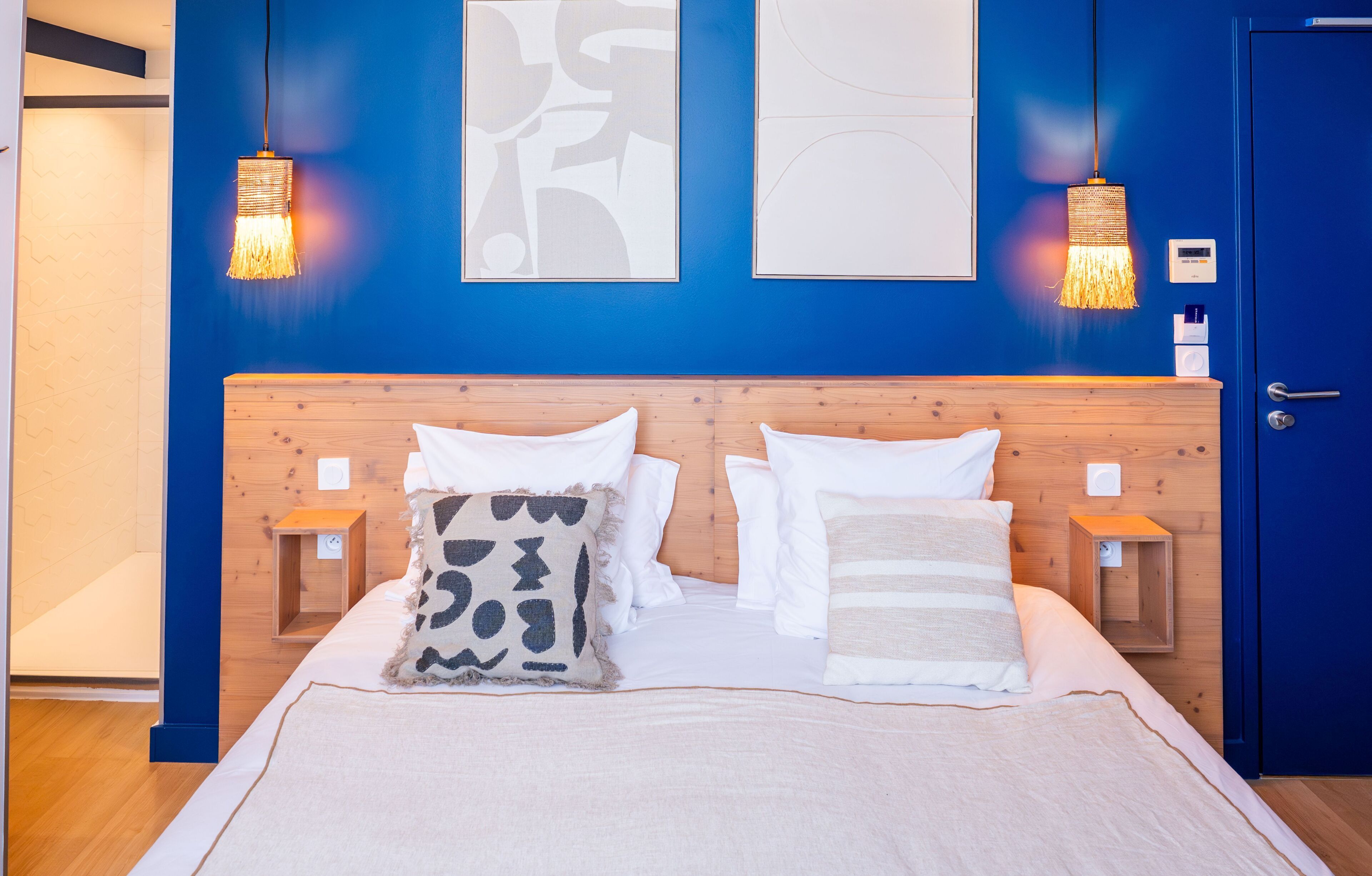 Una habitación de hotel con cabecera de madera, dos mesitas de noche, una cama con ropa de cama blanca y estampada, y dos lámparas fijadas a la pared.