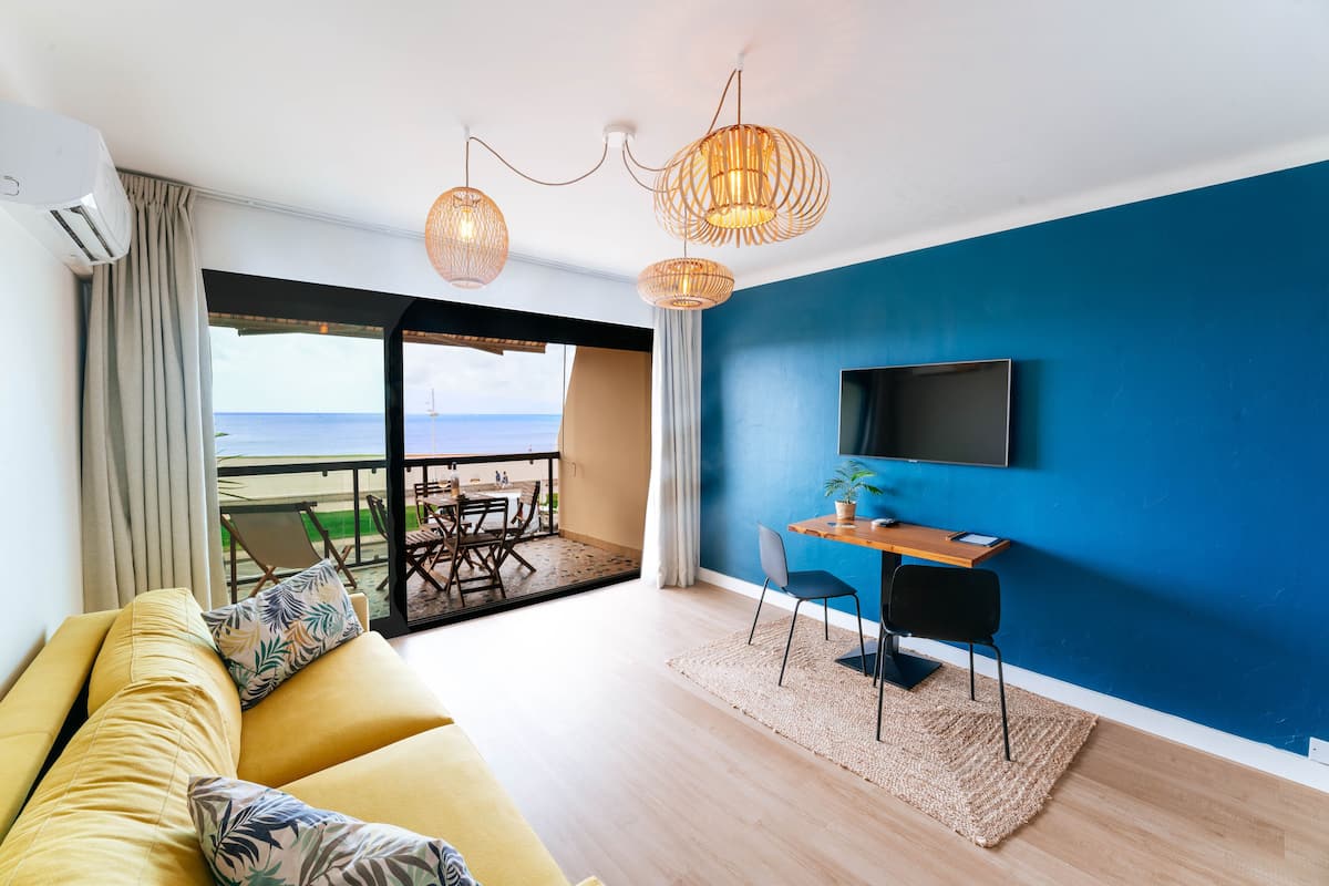 A modern living room with a sofa, a dining table, and a TV. There is a balcony with a view of the sea.