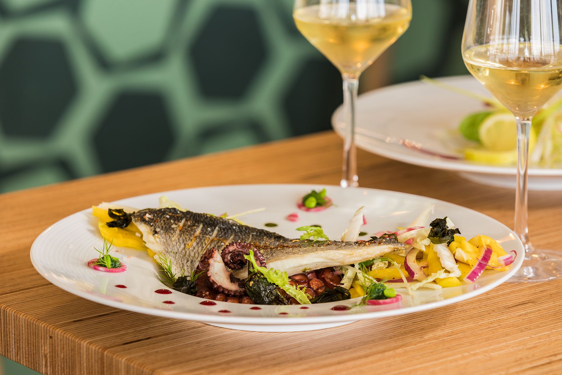 A plate of food with a fish and salad, accompanied by two glasses of white wine on a wooden table.