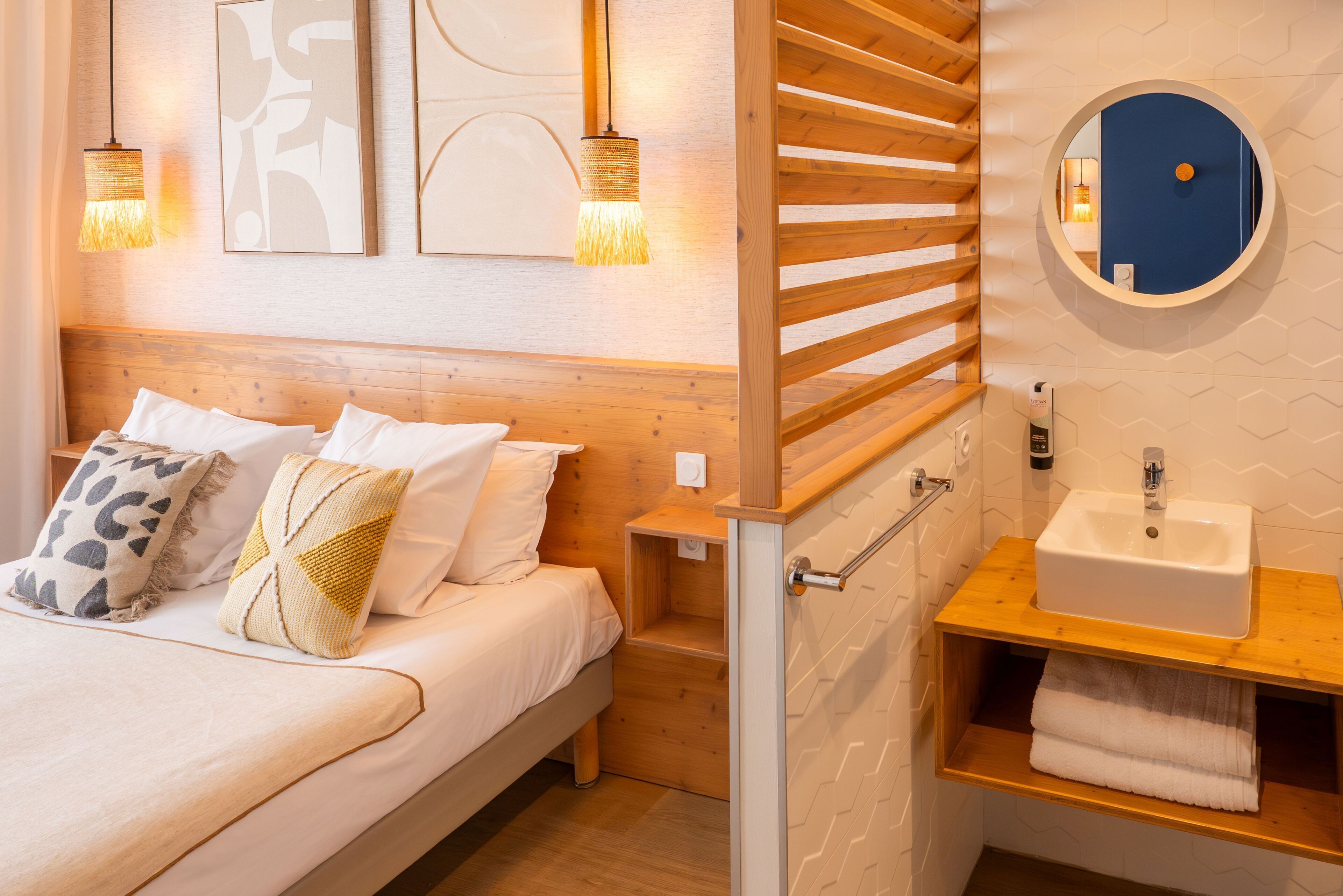Un dormitorio con cama, mesitas de noche, cabecera de madera, espejo redondo y baño con lavabo y toallero.