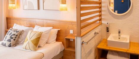 Un dormitorio con cama, mesitas de noche, cabecera de madera, espejo redondo y baño con lavabo y toallero.