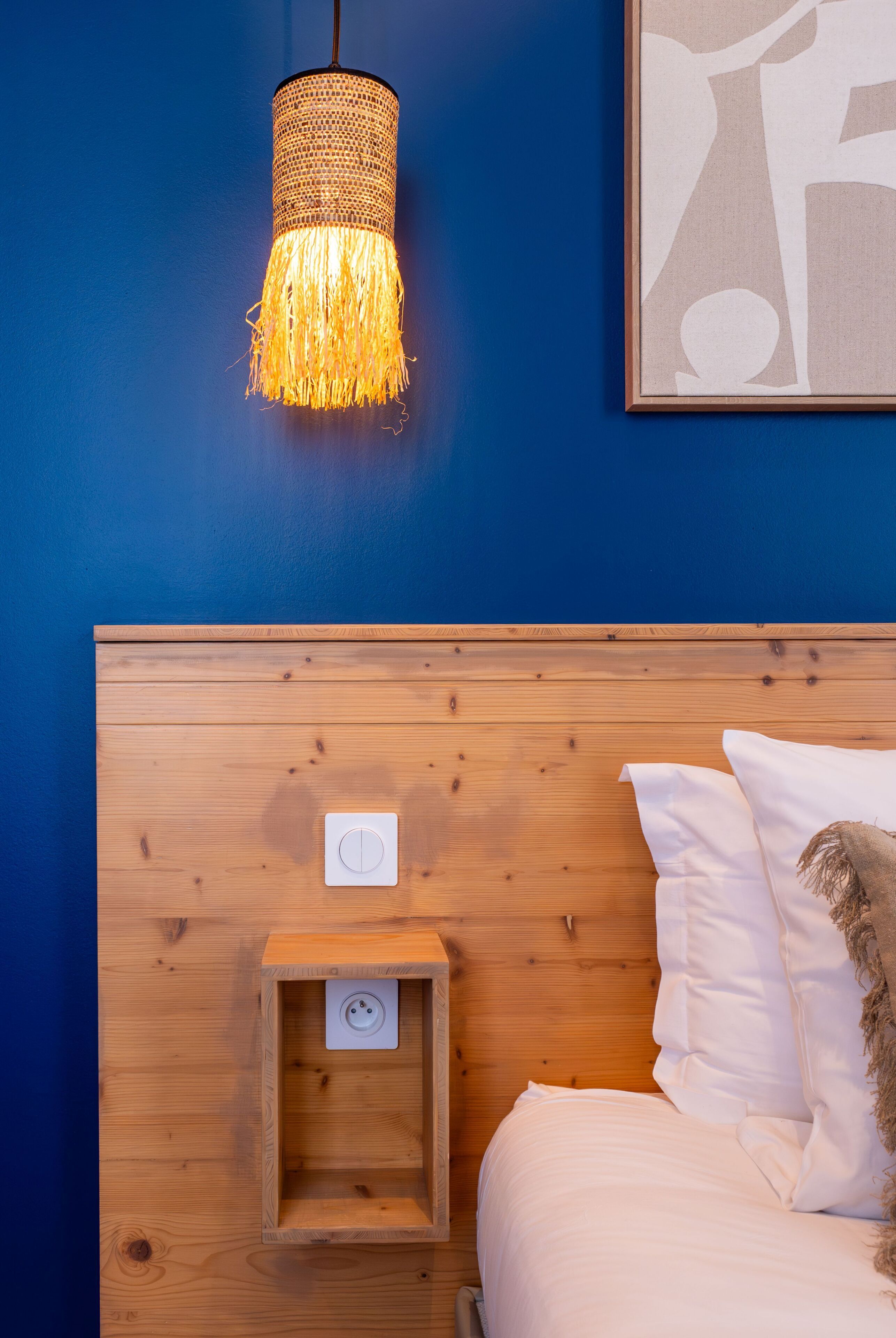 A bedroom with a wooden headboard, a bedside table with a power outlet, a hanging lamp, and a framed abstract artwork on the wall.