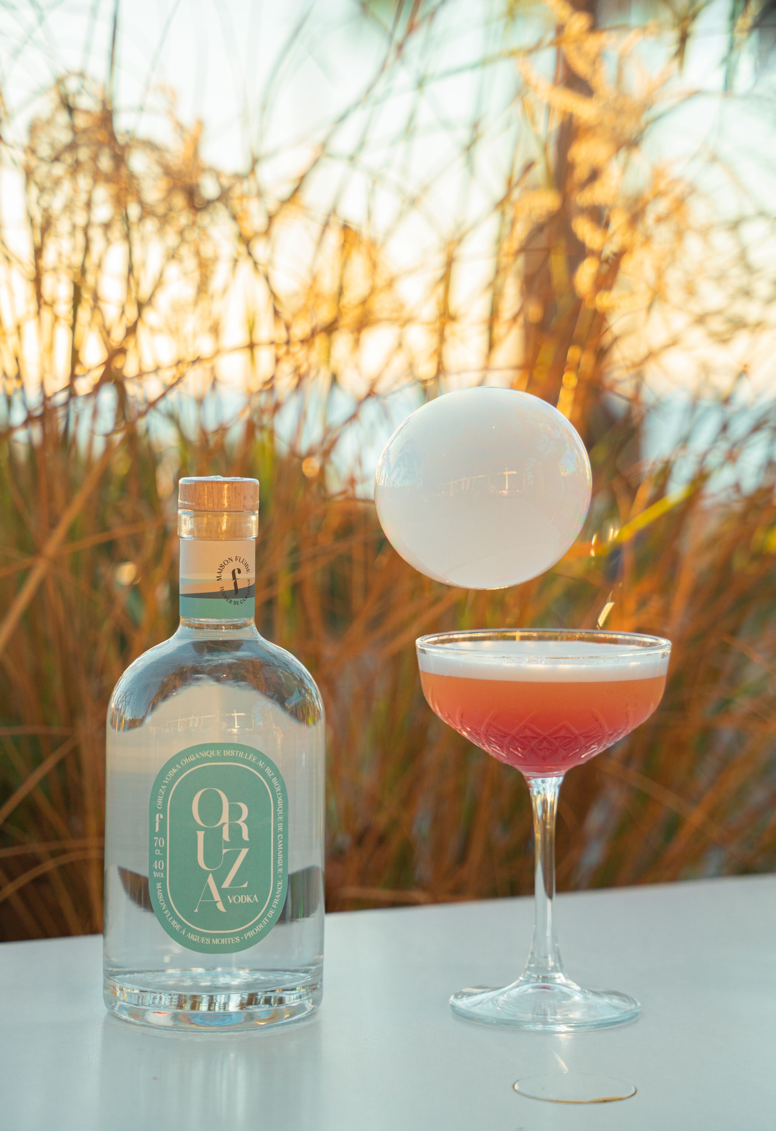 Una botella de vodka Oru y un cóctel sobre una mesa con un fondo natural desenfocado.