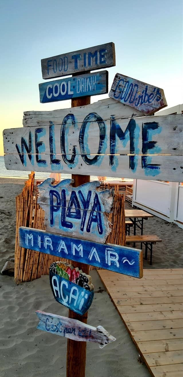 Um painel de madeira com vários indicadores de direção, incluindo “BEM-VINDO”, “PRAIA”, “MIRAMAR”, “HORÁRIO DE REFEIÇÕES”, “BEBIDA GELADA” e “BOA SORTE”.