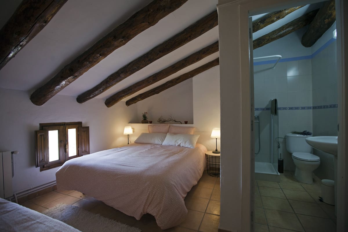 Une chambre avec un grand lit, des lampes de chevet, une fenêtre et une vue sur une salle de bain équipée de toilettes et d’un lavabo.