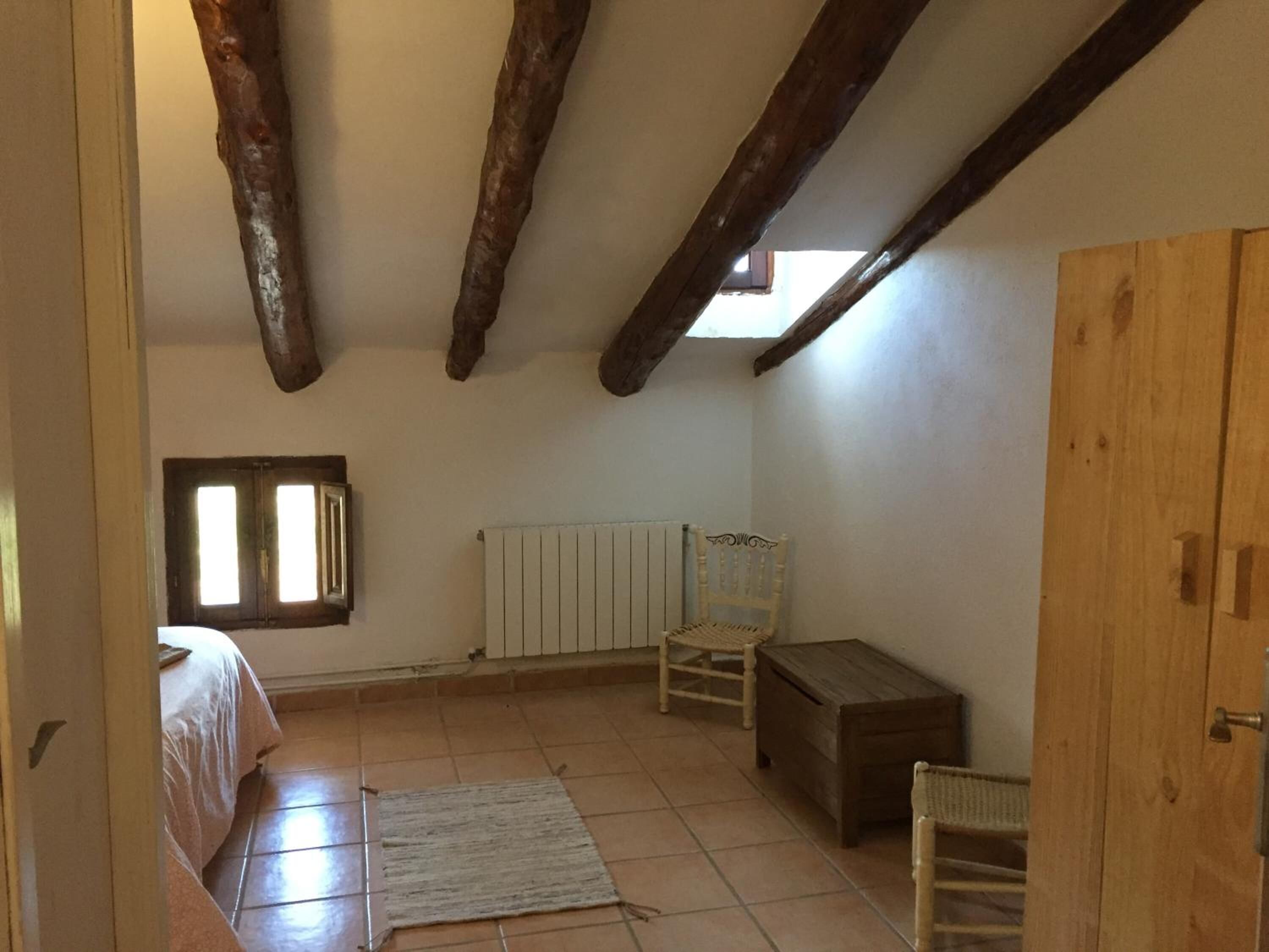 Una habitación con techo inclinado, vigas de madera, radiador, cama, silla, mesita y ventana.