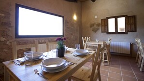 Comedor con mesa de madera dispuesta para cuatro, sillas, planta en maceta y ventana con contraventanas.