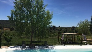 Una piscina con escalones de acceso, rodeada de césped, árboles y una pérgola con sillas.