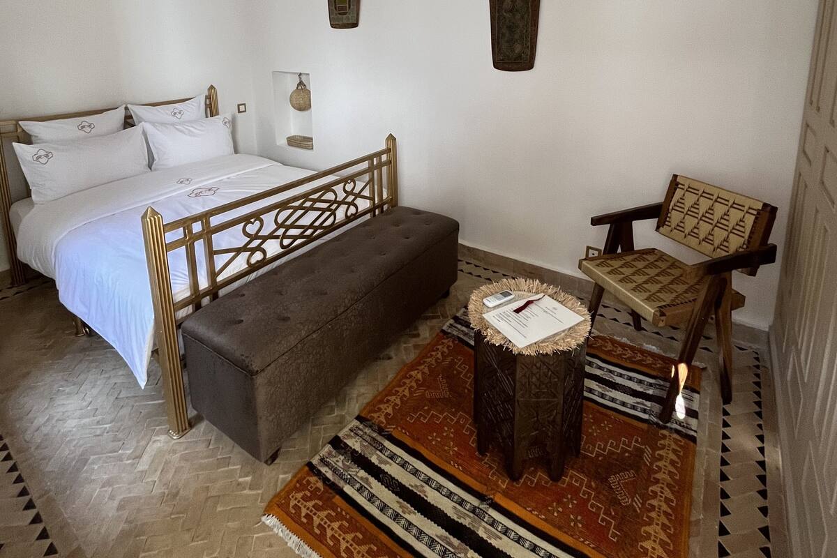 A bedroom with a bed, a bench, a chair, a small table, and a hanging light fixture.