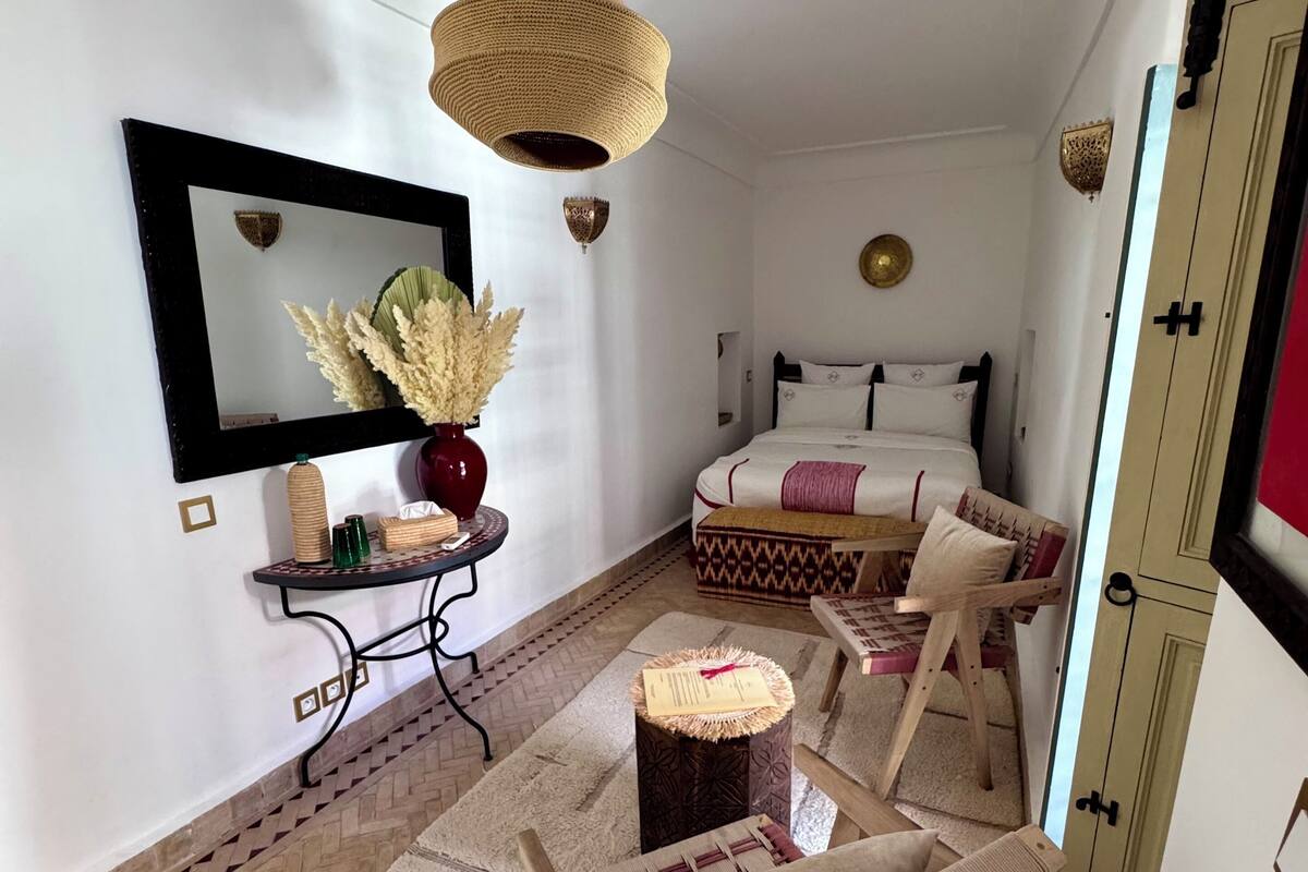 A cozy room with a bed, a chair, a small table, a mirror, and a hanging light fixture.
