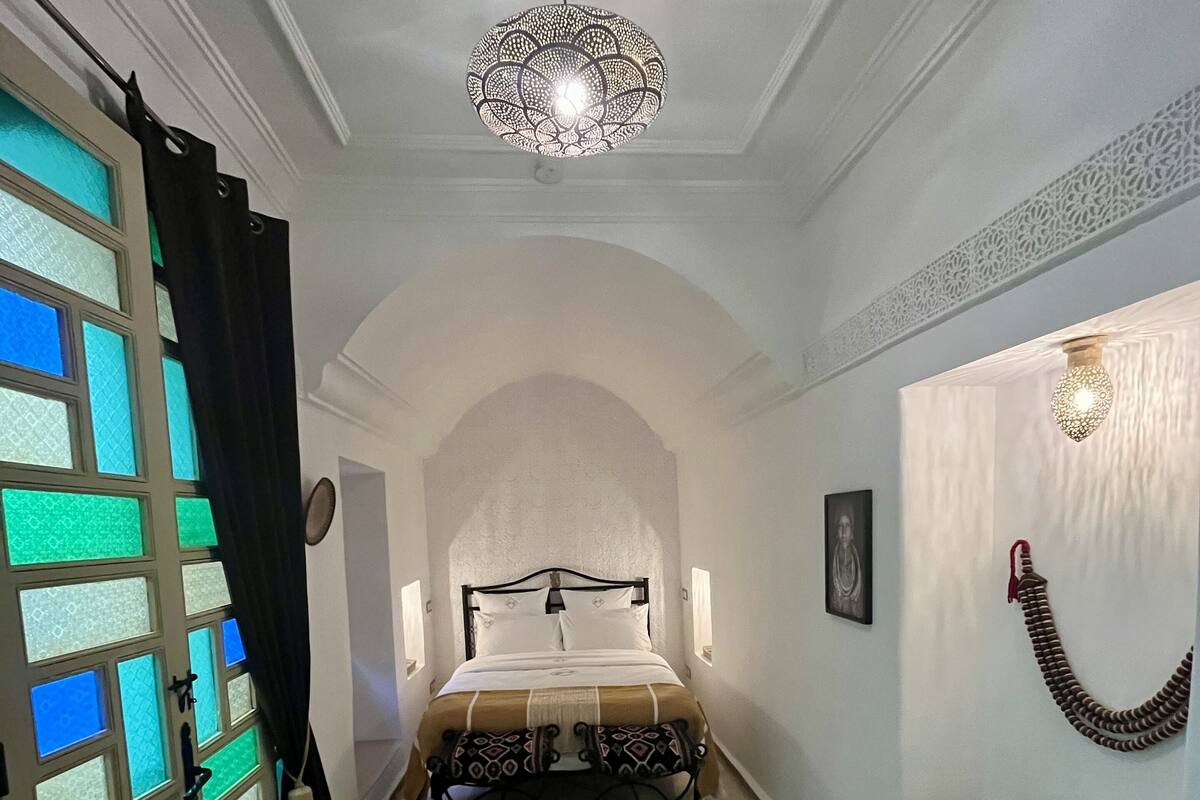 A bedroom with a bed, a chandelier, a patterned rug, and a decorative wall piece.