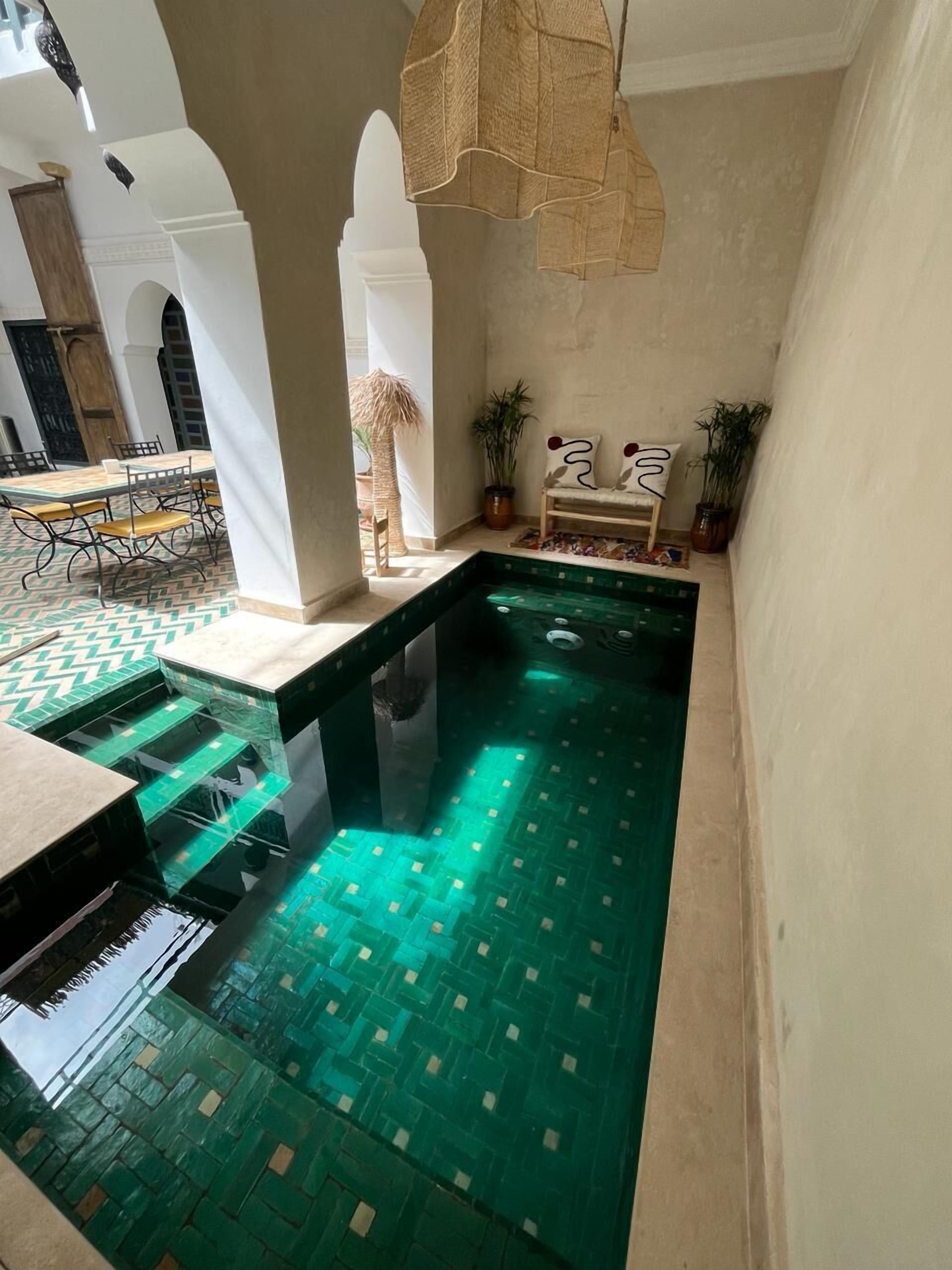 Una piscina con un diseño geométrico de azulejos, un comedor con mesa y sillas, y un espacio de descanso con un banco y plantas en macetas.
