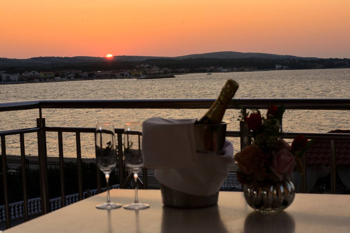 A table with a bottle of champagne, a glass, and a vase with flowers, overlooking a body of water with a sunset in the background.