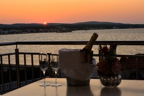 A table with a bottle of champagne, a glass, and a vase with flowers, overlooking a body of water with a sunset in the background. - Sunset Apartments (Vir)
