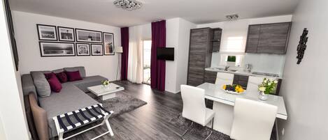 Un apartamento moderno con un sofá gris, una mesa de comedor con sillas, una zona de cocina y una sala de estar con televisor.