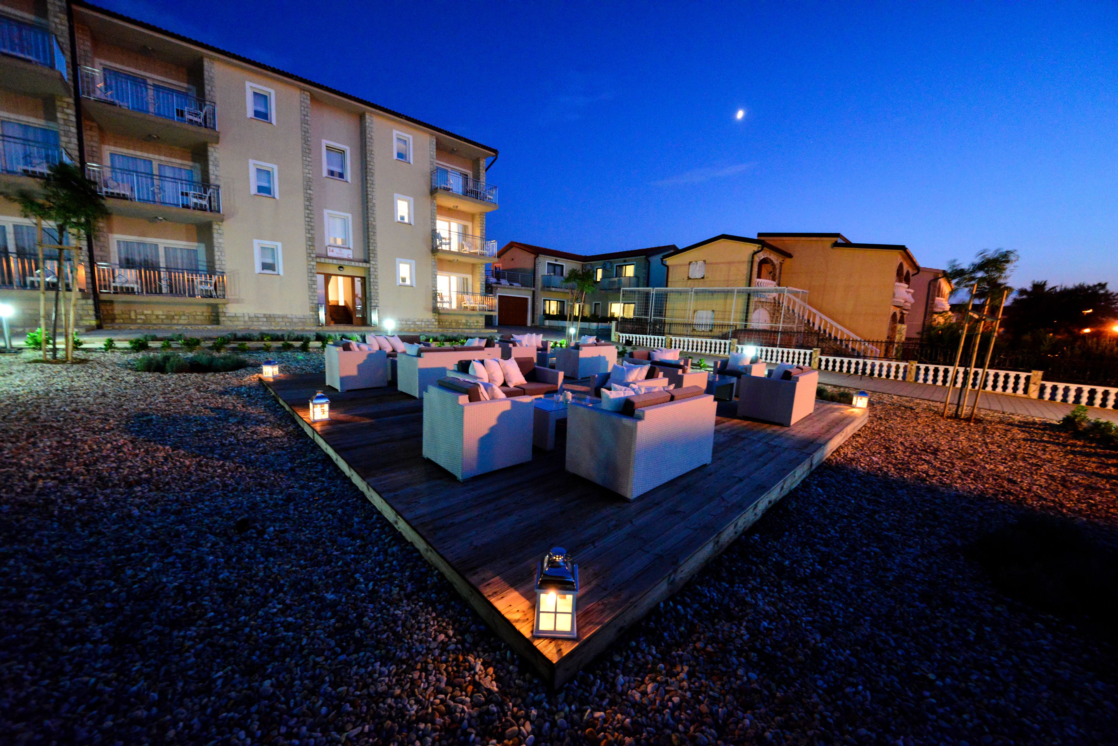 An outdoor lounge area with sofas and tables, surrounded by a wooden deck and gravel, with buildings in the background.