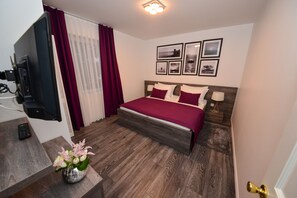 A bedroom with a bed, a TV, a vase with flowers, and framed pictures on the wall. - Sunset Apartments (Vir)