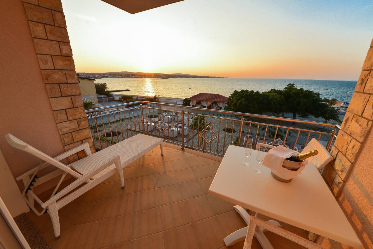 A balcony with a view of the sea at sunset, a table with a bottle of champagne, and two white lounge chairs.