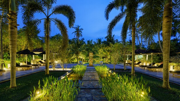 A pathway flanked by palm trees and illuminated by ambient lighting, leading to a seating area with umbrellas. - JH Villa (Unawatuna)