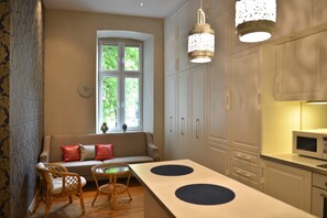 A modern kitchen with white cabinets, a microwave, a dining table with two chairs, and a window with a view of greenery. - La Gioia Magic Orient Apartments (Kraków)