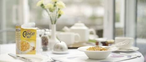 A breakfast setup with a bowl of cereal, a teapot, cups, and a box of cereal on a table with a view of the outdoors.