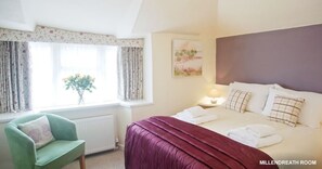 A hotel room with a bed, a green armchair, a window with curtains, a bedside table with a lamp, and a painting on the wall. - The Old Bridge House (Looe)