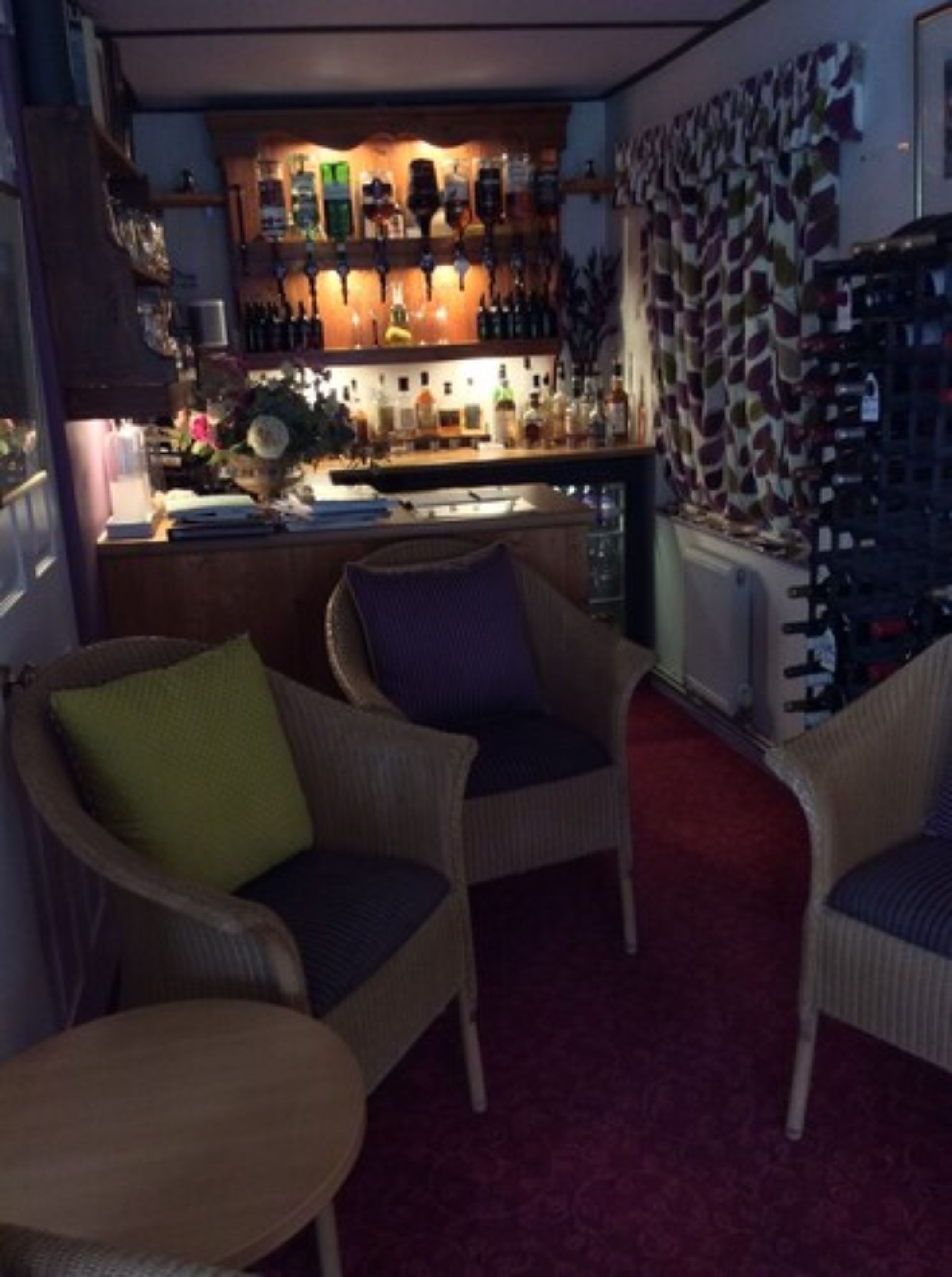 A bar area with a variety of bottles and a wall of wine racks. There are wicker chairs and a small table in the foreground.