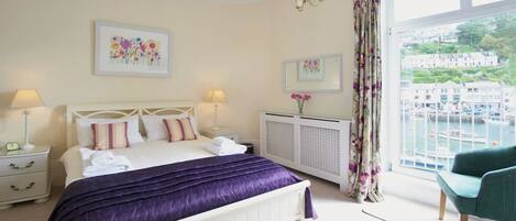 A bedroom with a bed, two bedside tables, a radiator, a window with curtains, and a view of buildings and water.