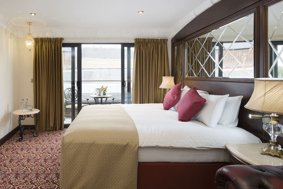 A hotel room with a large bed, a small table with a lamp, and a view of the outside through a large window.