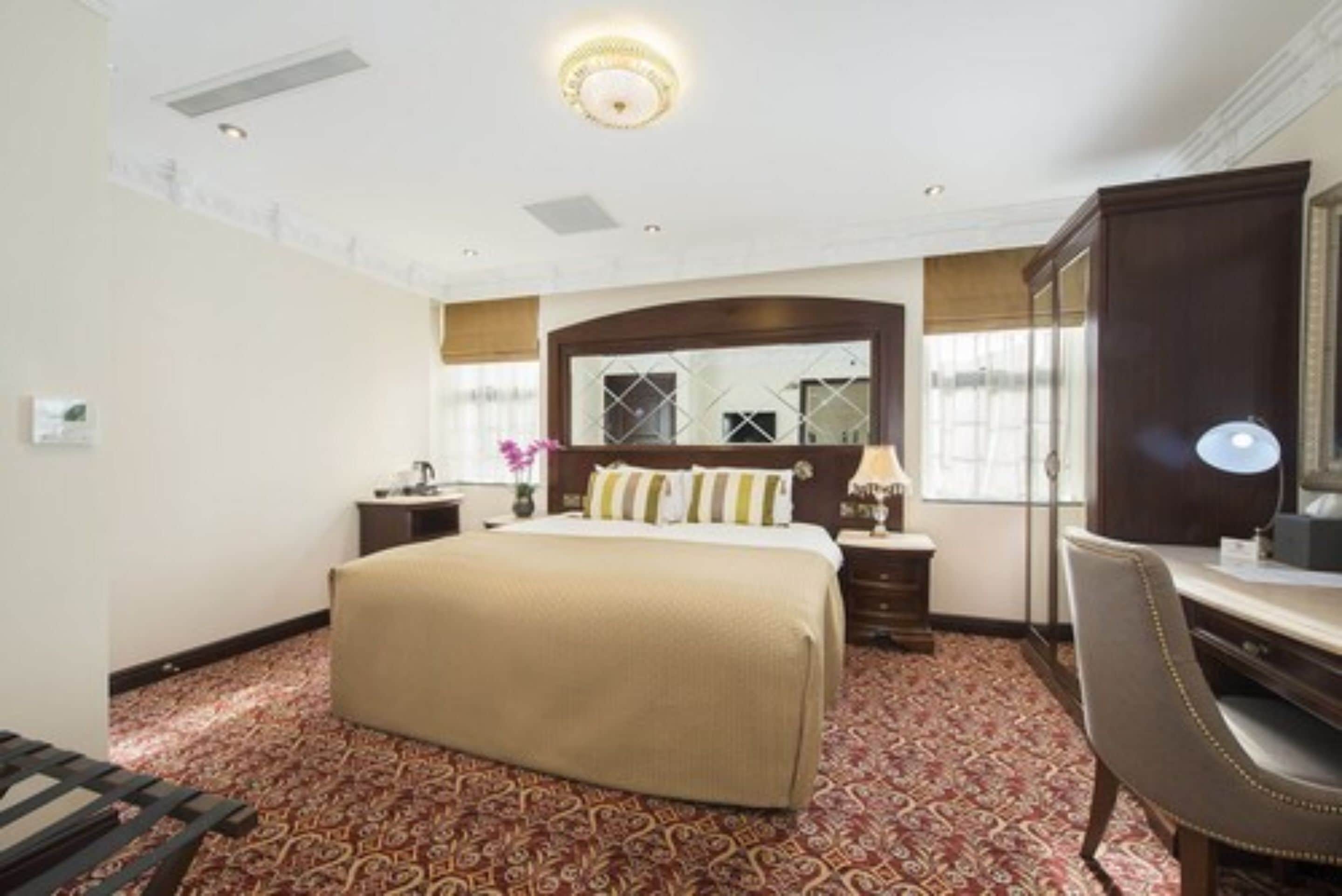 Habitación de hotel con una cama grande, un escritorio con silla, un espejo y una alfombra estampada.