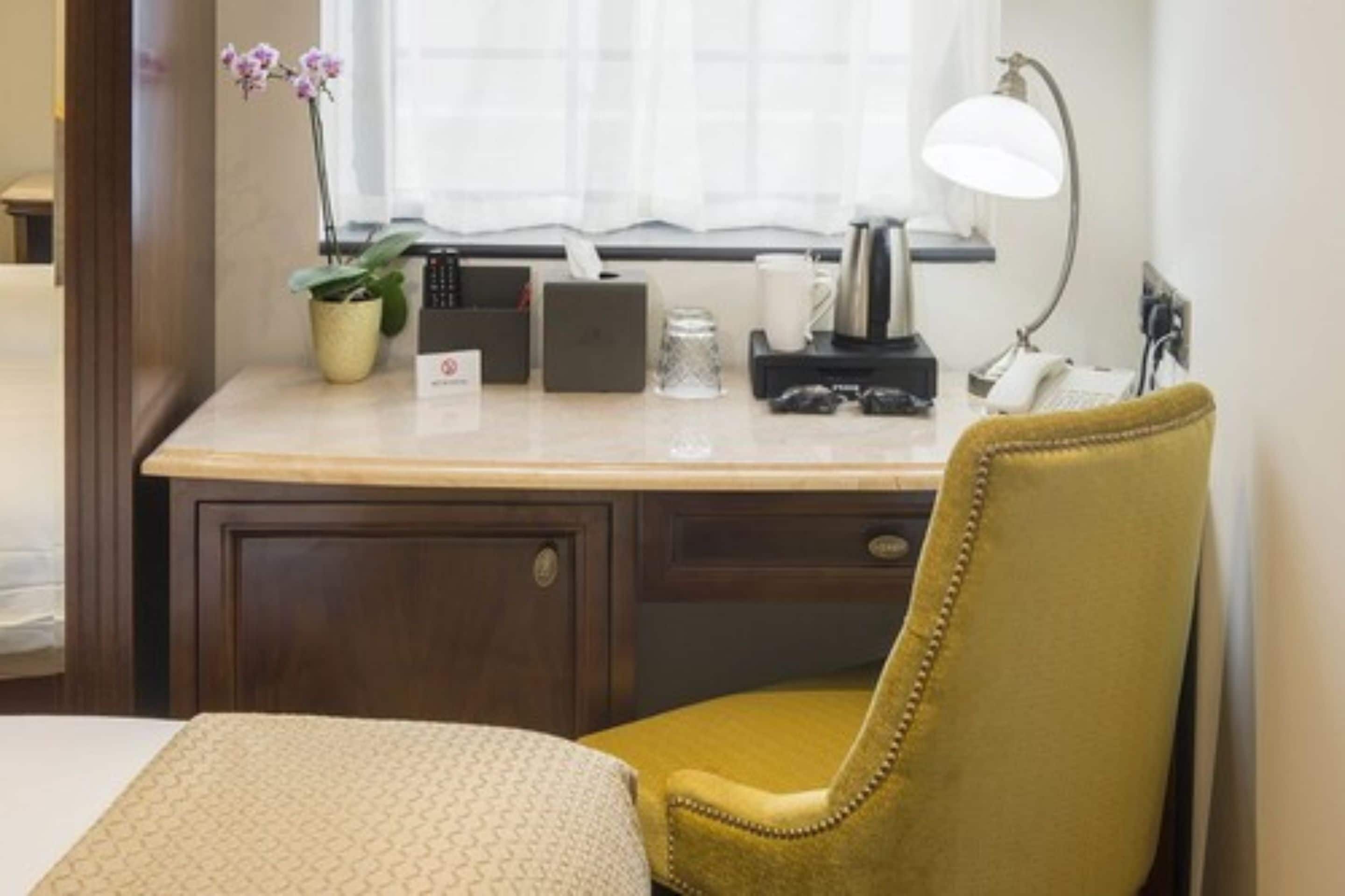 Une chambre d’hôtel équipée d’un bureau, d’une chaise, d’une fenêtre avec des rideaux, d’une lampe, d’un téléphone, d’une machine à café, d’une plante et d’une boîte de mouchoirs.